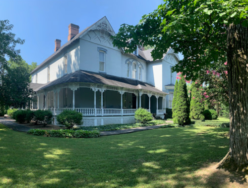 A large, historic house with a wraparound porch, surrounded by lush greenery and trees on a sunny day.