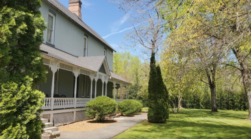 A charming historic house with a porch, surrounded by lush greenery and trees under a clear blue sky.