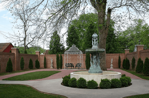 A serene garden with a fountain, gazebo, and manicured hedges surrounded by brick walls and trees.