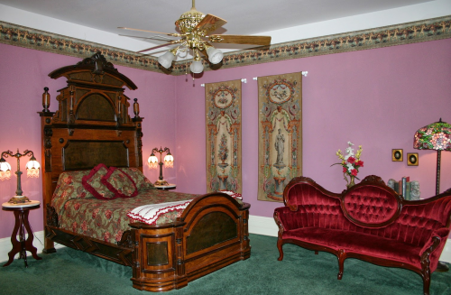 A vintage bedroom with a wooden bed, ornate lamps, a red velvet sofa, and floral decor on pink walls.