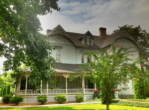 A large, historic house with a wraparound porch, surrounded by greenery and trees under a cloudy sky.
