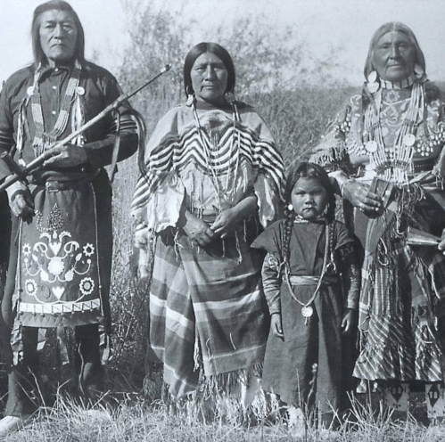 A historical black-and-white photo of four Indigenous individuals in traditional attire, standing together outdoors.