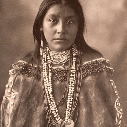 A portrait of a Native American woman wearing traditional attire and multiple strands of beads and necklaces.