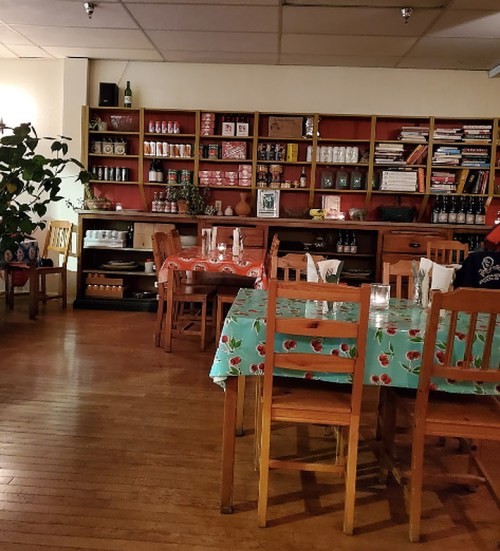 Cozy restaurant interior with wooden tables, colorful tablecloths, and shelves filled with jars and books in the background.
