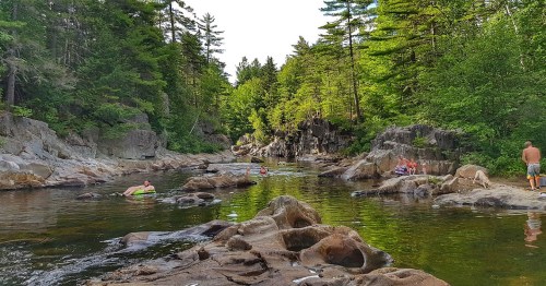 A serene river scene with people swimming and relaxing among rocks, surrounded by lush green trees.