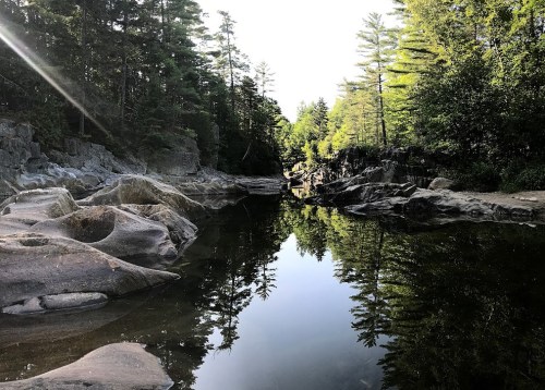 A serene landscape featuring a calm river reflecting trees and rocks, surrounded by lush greenery and sunlight.