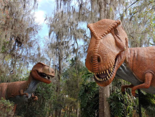Two large dinosaur sculptures in a lush, green forest with Spanish moss hanging from trees.