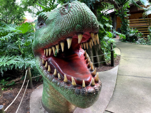 A large, colorful dinosaur sculpture with an open mouth, surrounded by lush greenery in a park setting.