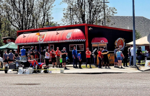 A busy burger restaurant with a red roof, featuring a long line of people waiting outside on a sunny day.