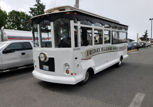 A white trolley bus with "Friday Harbor Boat Tours" signage parked in a lot, surrounded by vehicles and trees.