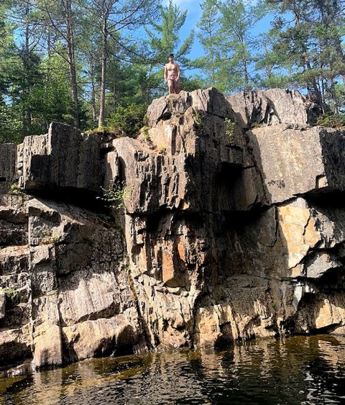 A person stands on a rocky cliff above a calm body of water, surrounded by trees under a clear blue sky.