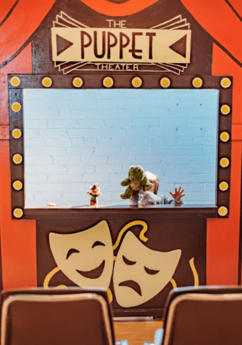 A colorful puppet theater stage with puppets displayed, featuring a frog and a flower, set against a bright backdrop.