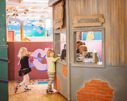 Children interacting at a playful bank-themed area, with a withdrawal window and colorful decor in the background.
