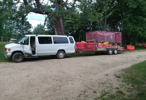 A white van parked on a dirt road, towing a trailer with colorful kayaks and a pink canopy. Trees are in the background.
