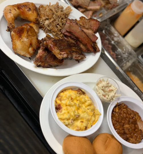 Two plates of barbecue featuring chicken, pulled pork, ribs, coleslaw, baked beans, macaroni and cheese, and rolls.