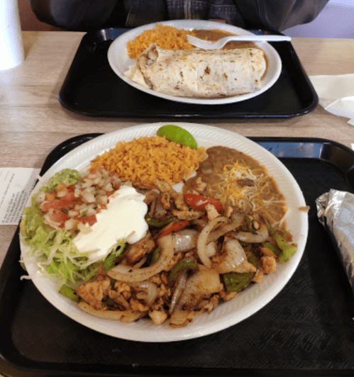 A plate of grilled chicken with peppers, onions, rice, beans, guacamole, and sour cream, with a burrito in the background.