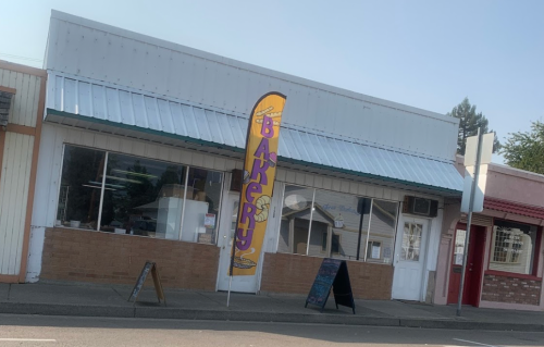 A bakery storefront with a colorful sign and large windows, featuring a striped awning and sidewalk chalkboards.