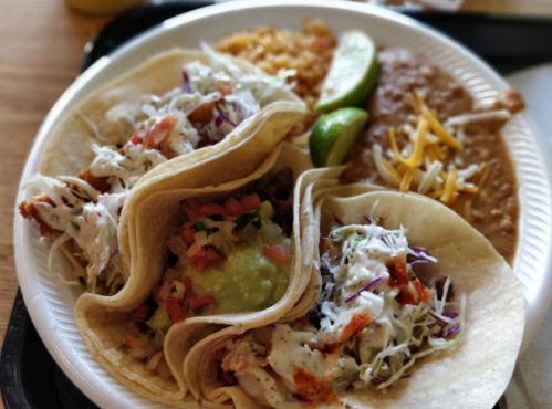 A plate with three tacos filled with chicken, guacamole, and toppings, served with rice and beans on the side.