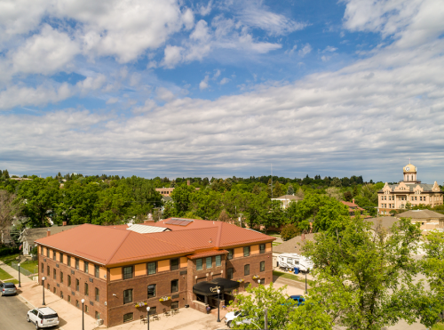 Aerial view of a brick building with a red roof, surrounded by trees and a cloudy sky, with a historic building in the background.
