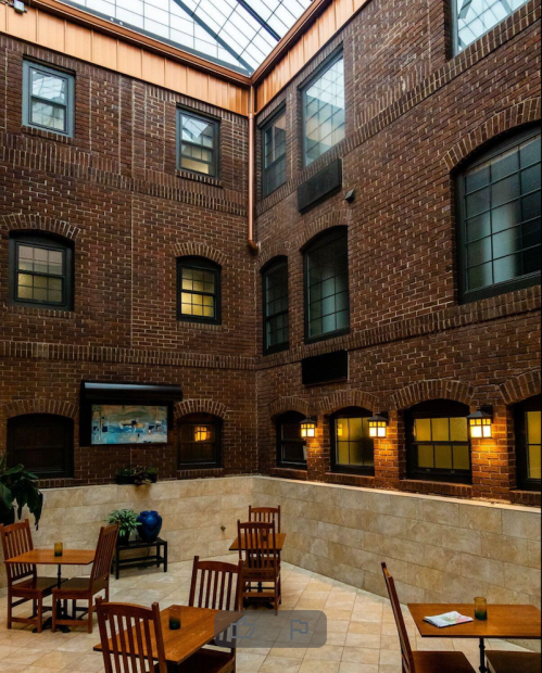 Interior view of a brick atrium with wooden tables and chairs, featuring large windows and a glass roof.