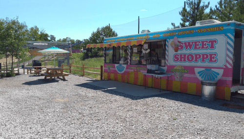 A colorful food truck named "Sweet Shoppe" with outdoor seating, surrounded by trees and gravel.