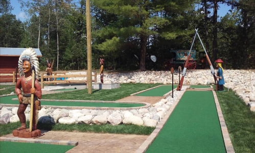 A mini-golf course featuring stone sculptures, green turf, and trees in a sunny outdoor setting.