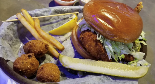 A crispy chicken sandwich with lettuce, pickles, fries, and hushpuppies on a plate, accompanied by dipping sauce.