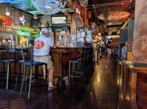 A lively bar interior with wooden decor, patrons seated at the counter, and colorful lights illuminating the space.