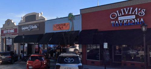 A row of storefronts featuring Liberty & Goods, Oyster 84, and Olivia's Taverna under a clear blue sky.