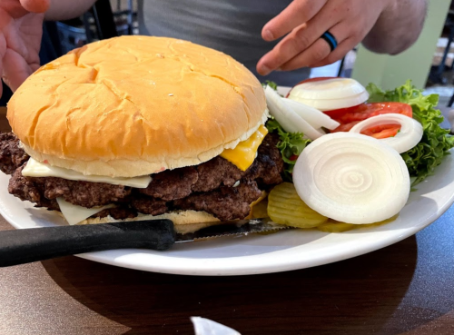 A large burger with multiple beef patties, cheese, lettuce, tomato, onions, and pickles on a plate, with a knife beside it.