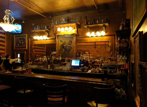 A vintage bar interior with a wooden counter, shelves of liquor, and a large portrait of a musician on the wall.