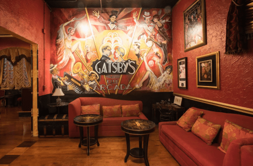 A cozy lounge with red walls, featuring a vibrant mural of Gatsby's, and stylish seating arrangements.