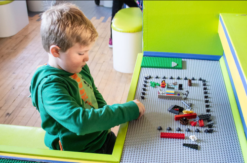 A young child builds with colorful LEGO bricks on a gray baseplate in a bright, playful environment.