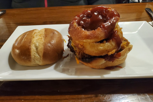 A burger with onion rings and barbecue sauce, served on a white plate next to a plain bun.
