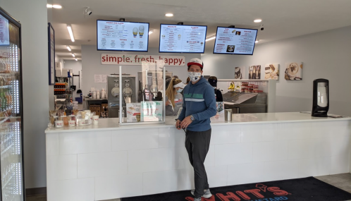 A man wearing a mask stands at the counter of an ice cream shop, with menus displayed on screens above.