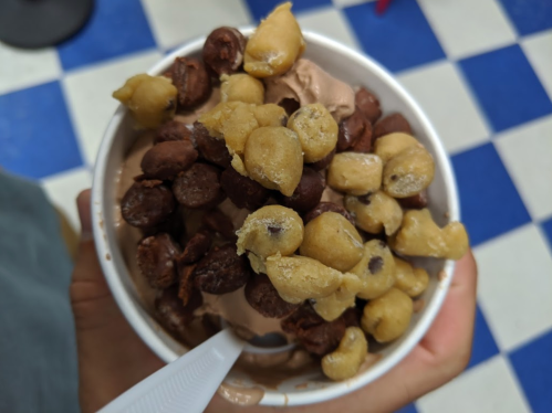 A cup of chocolate ice cream topped with cookie dough bites and chocolate-covered snacks, held in a hand.