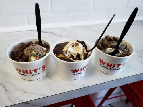 Three cups of frozen custard with toppings, including sprinkles and chocolate sauce, on a marble table.