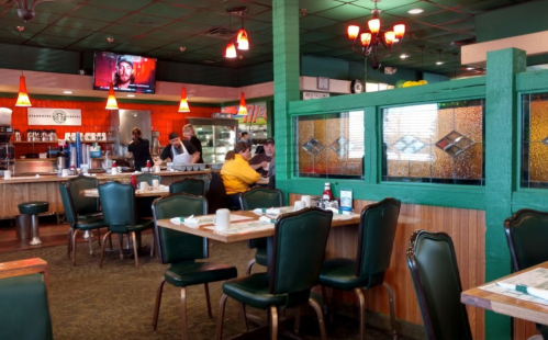 A cozy diner interior with green booths, tables set for dining, and a TV showing a program in the background.