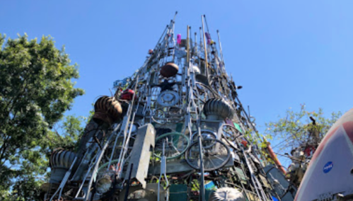 A towering sculpture made of metal and various objects, reaching towards a clear blue sky, surrounded by trees.