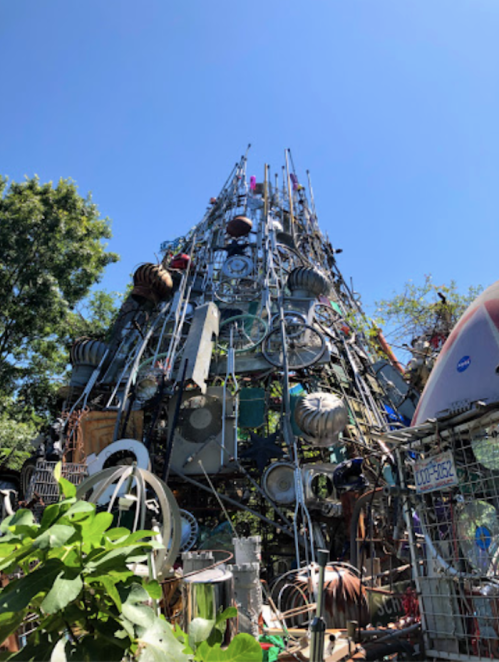 A towering sculpture made of various metal objects and debris, set against a clear blue sky and green trees.