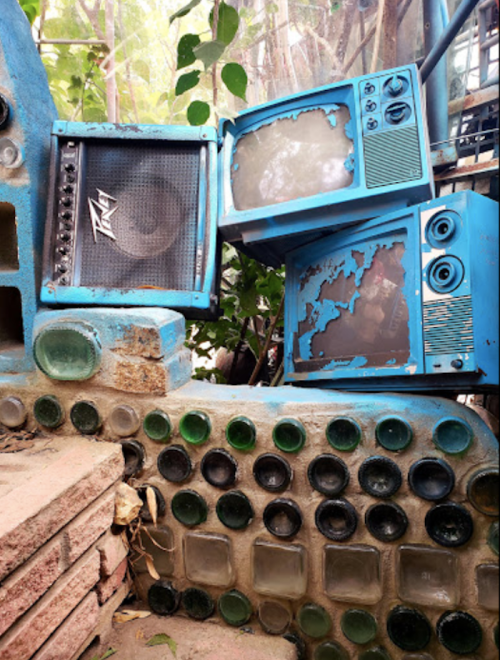 Two vintage blue televisions and a speaker are stacked against a wall made of colorful glass bottles and bricks.