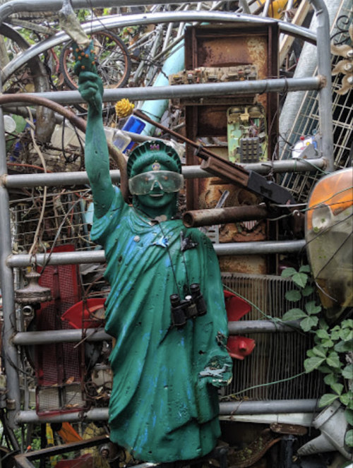 A weathered statue of the Statue of Liberty amidst a cluttered junkyard, wearing goggles and holding binoculars.