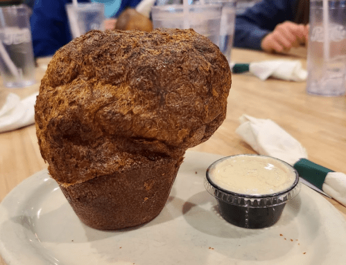 A large, golden-brown muffin on a plate next to a small cup of creamy dipping sauce.