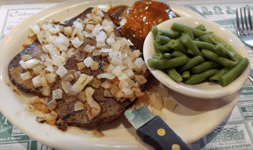 A plate of steak topped with onions, barbecue sauce, a side of green beans, and a knife on the side.