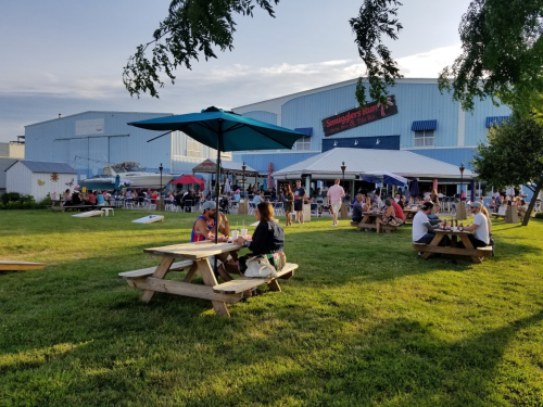 A lively outdoor scene with people dining at picnic tables under umbrellas, surrounded by a grassy area and a building.