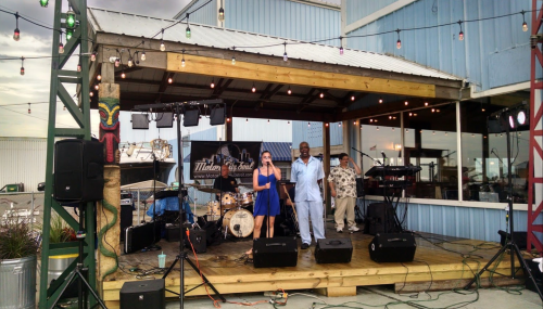 A woman and a man speak on stage at a lively outdoor venue with a band setup and string lights.