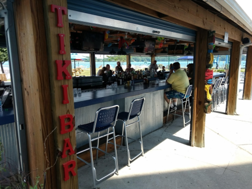 A tiki bar with a long counter, stools, and patrons enjoying drinks in a sunny outdoor setting.