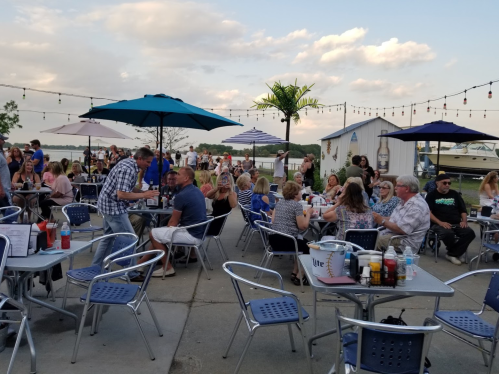 A lively outdoor dining area with people at tables, umbrellas, and a scenic view by the water.