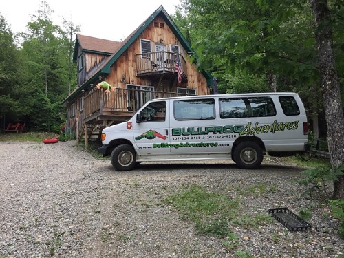 A white van with "Bullfrog Adventures" parked near a wooden cabin surrounded by trees and gravel.
