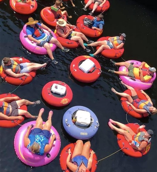 Aerial view of people relaxing on colorful inner tubes in a river, some chatting and enjoying the sun.